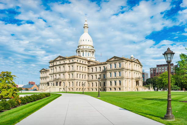 The Michigan State Capitol, a historic architectural landmark, stands prominently with the urban skyline of Lansing, Michigan, surrounded by green lawns and city buildings under an overcast sky.