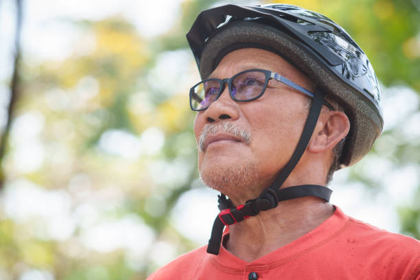 istock Asian Senior Cyclist man with a determined expression wearing eyeglasses and a protective helmet, looking up outdoors, dressed in an orange shirt, enjoying a sunny day in nature 2184283607