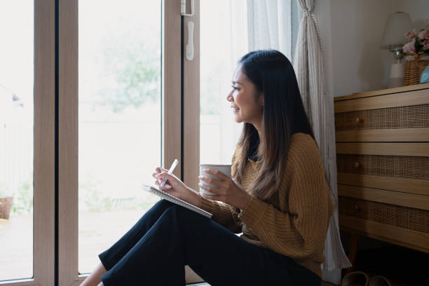 istock Beautiful young woman writing in notebook while looking out the window 2184289166