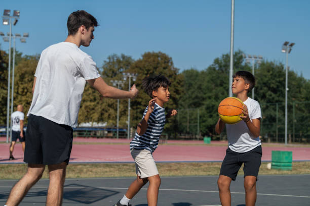 istock Boys engage in outdoor basketball training with a coach, practicing dribbling, passing, and shooting skills on a court surrounded by greenery under bright natural light, fostering teamwork and coordination. 2184292237