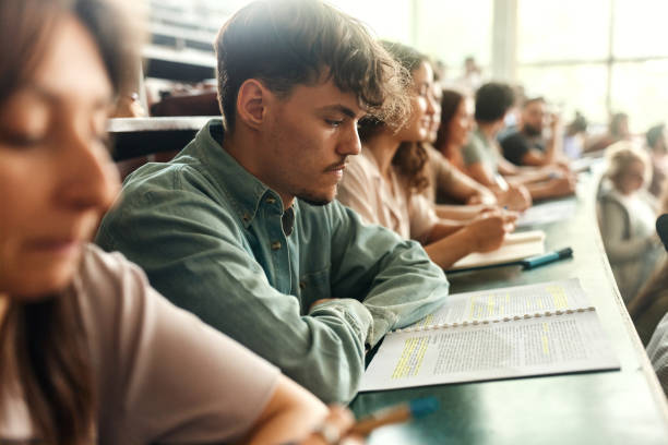 istock Young male student reading textbook on a class in lecture hall. 2184293232