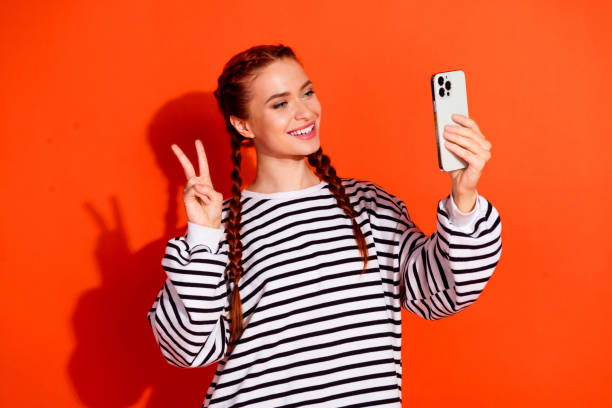 istock Joyful young woman in casual striped pullover taking selfie against vibrant orange background 2184303501