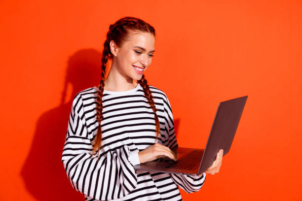 istock Young female with red hair and striped pullover using laptop against vibrant orange background 2184303555