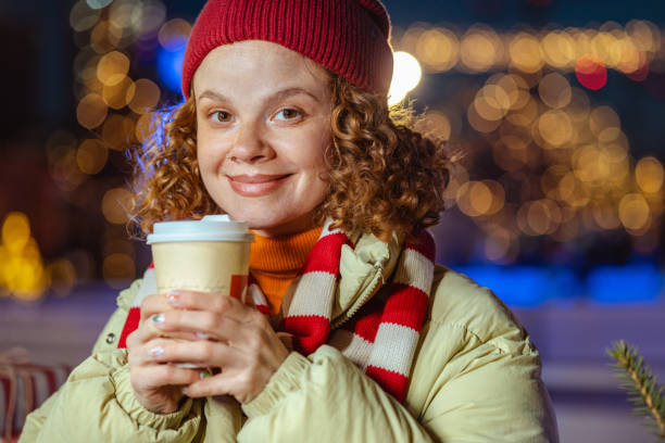 A woman in a cozy winter outfit enjoys a warm drink, smiling with closed eyes against a backdrop of festive holiday lights. The scene captures the essence of winter warmth and holiday joy.