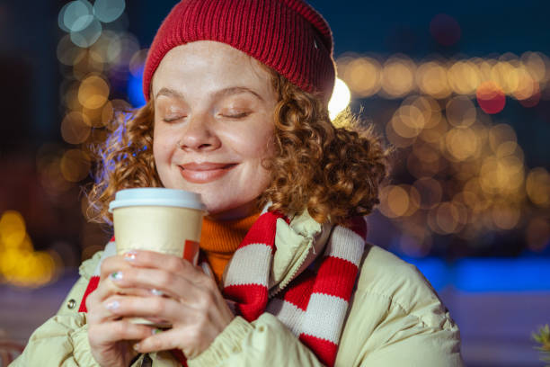 A woman in a cozy winter outfit enjoys a warm drink, smiling with closed eyes against a backdrop of festive holiday lights. The scene captures the essence of winter warmth and holiday joy.