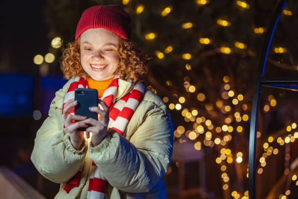 istock Smiling woman enjoying festive lights while using smartphone outdoors 2184307544