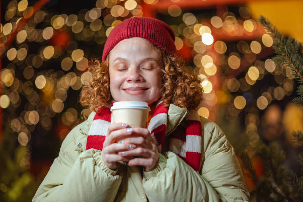 A woman in a cozy winter outfit holds a hot drink with a delighted expression. Sparkling holiday lights create a festive atmosphere, embodying warmth and happiness during the winter season outdoors.