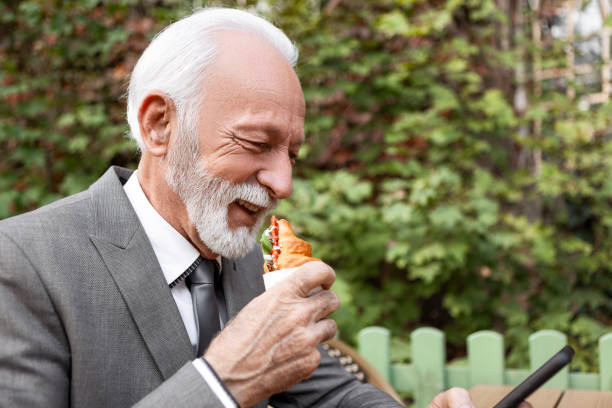 Senior Man Relaxing at Cafe with Coffee and Phone in Hand Eating Sandwich