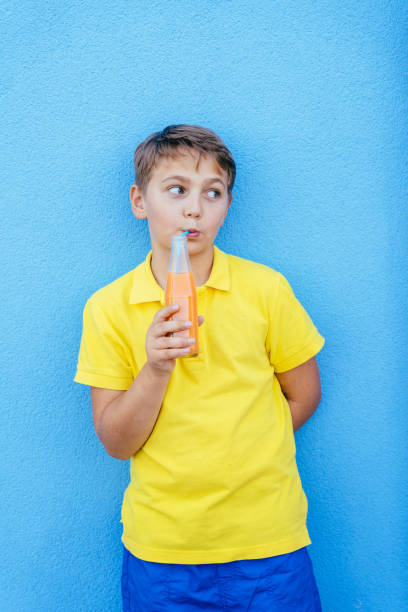 Young boy drinking soda stands against vibrant blue wall, wearing bright yellow shirt and blue shorts. Childhood with cheerful expression, playful, carefree childhood experiences.