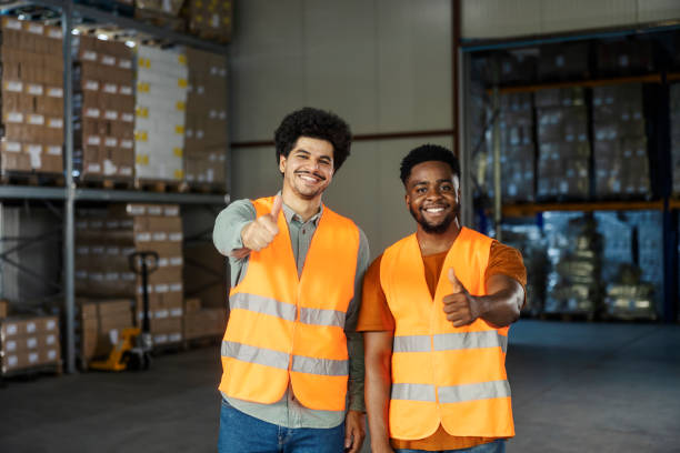 istock Two hardworking dedicated multicultural migrant blue collar storage workers giving thumbs up at camera. 2184333455