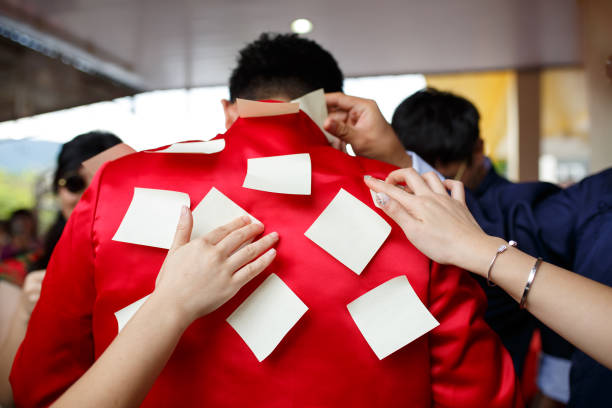 Man in a red shirt with several paper stickers being placed on his back by multiple hands. Groom gate crush game. Traditional Chinese wedding concept.