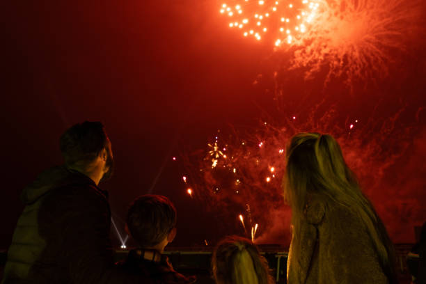Rear view shot of a family visiting an outdoor winter fireworks event. They are dressed warmly, standing in front of the firework lit sky in the North East of England. They are standing side by side looking up at the display.