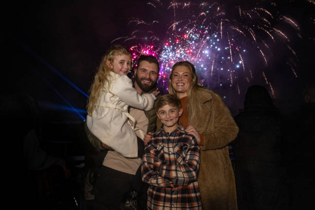 Family visiting an outdoor winter fireworks event. They are dressed warmly, standing in front of the firework lit sky in the North East of England. The father is carrying his daughter while they all stand with their arms around one another, smiling at the camera.\n\nVideos are available similar to this scenario.