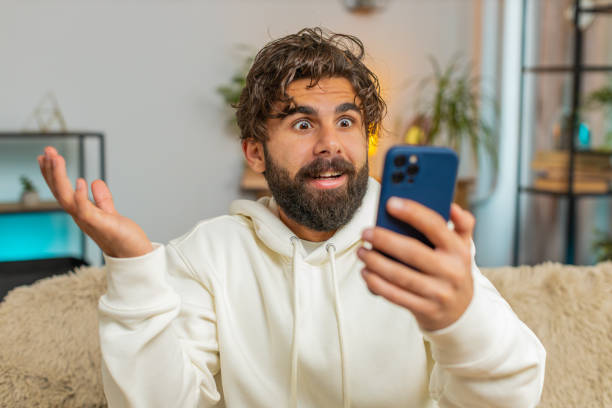 istock Surprised Indian bearded man using cellphone winning lottery sitting on sofa couch at home apartment 2184362450