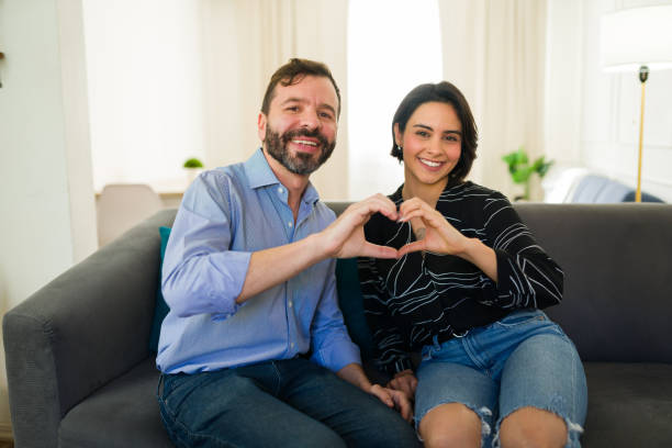 istock Smiling hispanic couple making heart sign with hands while sitting together on sofa, celebrating valentine's day at home 2184368969