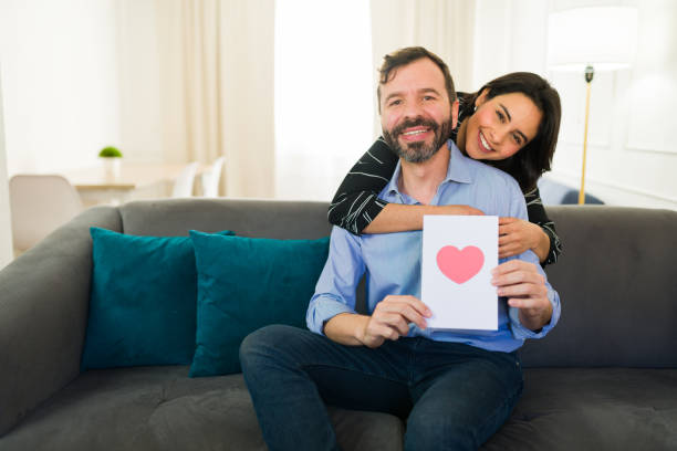 istock Couple sharing love and joy while sitting on the sofa at home, smiling and displaying a valentine's day card, celebrating with hearts and romance 2184369050