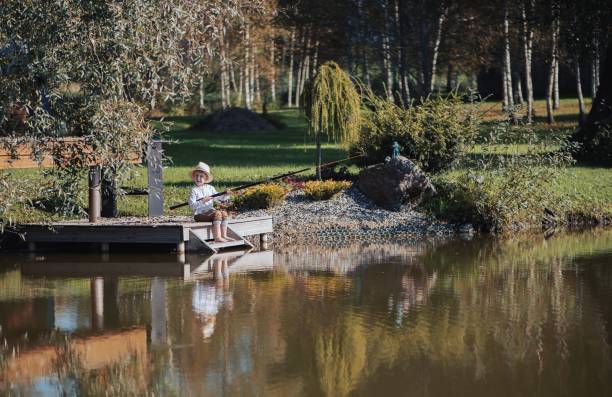 A young boy fishing on a tranquil pond dock surrounded by lush greenery and reflections in a serene outdoor setting.
