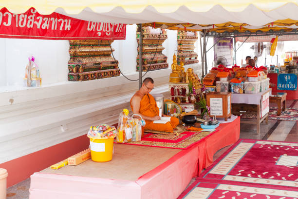 Thai monk sitting outside of tempe in Ayuthhaya . Monk is reading. He is sitting on a podest in aisle between buildings. People can purchuase gift baskets. Sceens is at Wat Kasattrathirat Worawihan in Ayuttaya. Monk is traditionally orange dressed. He is earing eyewear. At his right side is a donation box. Behind tgai men are sitting with many guft boxes and baskets.