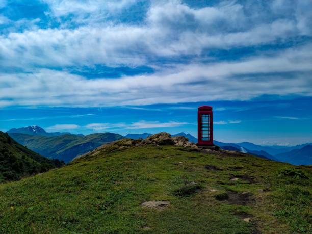 A red telephone booth stands on a grassy hill with a mountain backdrop under a vibrant blue sky.