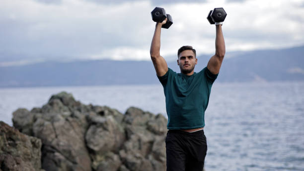 istock Young muscular man doing dumbbell shoulder press exercise on rocks at outdoor beachside 2184386910
