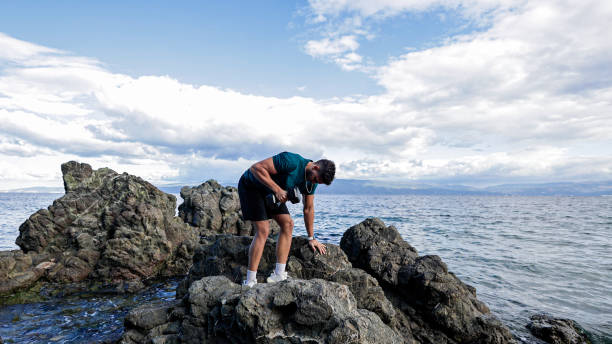 A young man performs a dumbbell tricep kickback exercise on rocky terrain by the sea. The natural outdoor setting emphasizes strength and fitness in a serene environment.