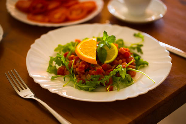 Elegant tuna tartare with fresh arugula, mint, and citrus accents on a white plate, centered on a wooden table, the background subtly showing additional breakfast dishes.