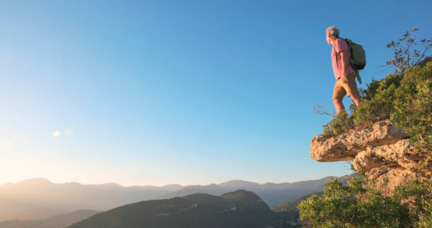 Mature man hikes onto cliff edge above mountains at sunset, Liguria