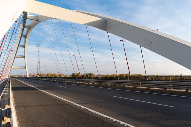 A road view of the Pentele Bridge, highlighting its distinctive arch structure and the array of red lamp posts and cables against a partly cloudy sky. The empty road and clear details emphasize the bridge's modern engineering and architectural elegance.