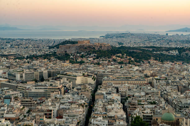 Elevated view highlights Athens' sprawling cityscape, with the iconic Acropolis illuminated against the evening sky, showcasing the blend of ancient and modern urban architecture.