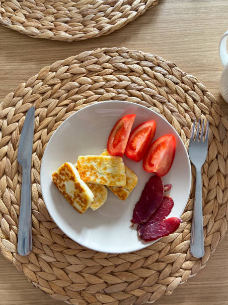 Cozy breakfast setting features plates with cheese, tomatoes, and cured meat. Woven placemats and wooden table provide a warm, inviting ambiance