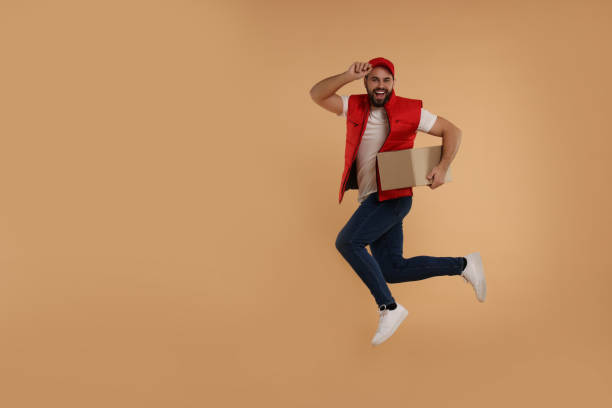 istock Happy young courier jumping with parcel on light brown background, space for text 2184421289