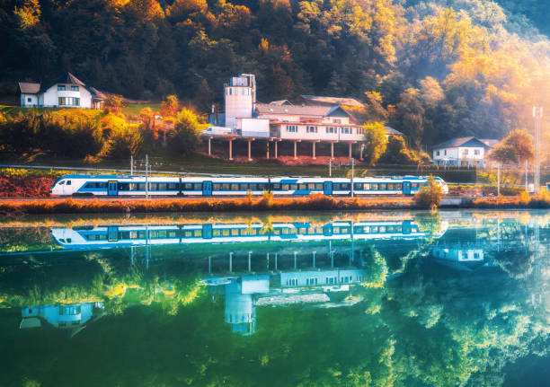 Beautiful blue modern high speed train and river in alpine mountains at sunrise in autumn. Passenger train, reflection in water, railroad, lake, orange trees in fall. Railway station in Slovenia