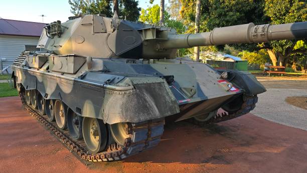 istock Military Leopard tank with a large cannon, camouflage paint, and tracks. It is displayed outdoors on a concrete surface, surrounded by trees and a building in the background. 2184430395
