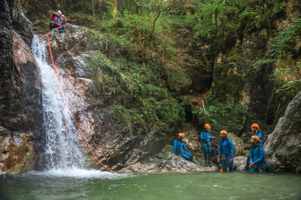 A group of diverse people, led by a skilled guide, slides down using rappel ropes, all adorned in neoprene suits and safety helmets during an exciting canyoning adventure.