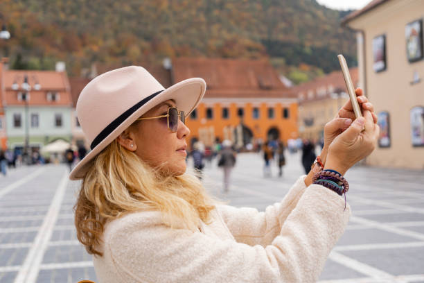 istock Woman with phone taking selfie in vibrant town square, dressed warmly with autumn backdrop. 2184453549