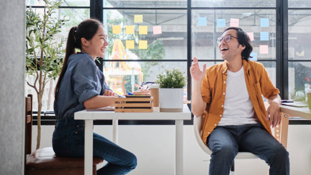 istock Two Asian workers at working desks near windows talk together. 2184460149