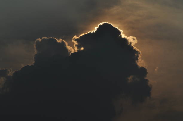 Dark silhouetted cloud with golden sunlight peeking from behind, creating a dramatic and atmospheric scene in the evening sky.