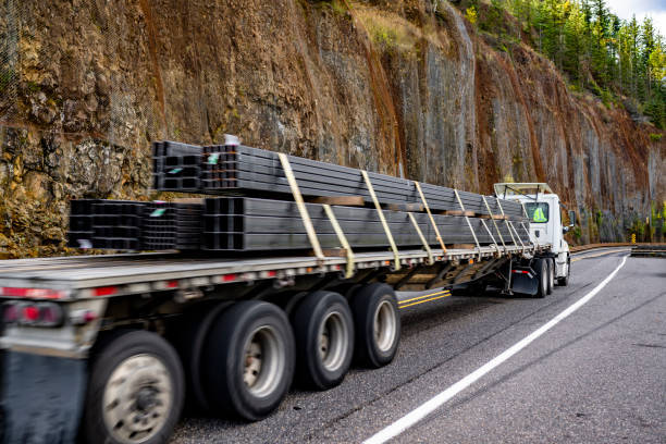 istock Day cab white big rig semi truck transporting heavy fastened metal cargo on flat bed semi trailer running on narrow mountain road with rock wall on the side 2184488924