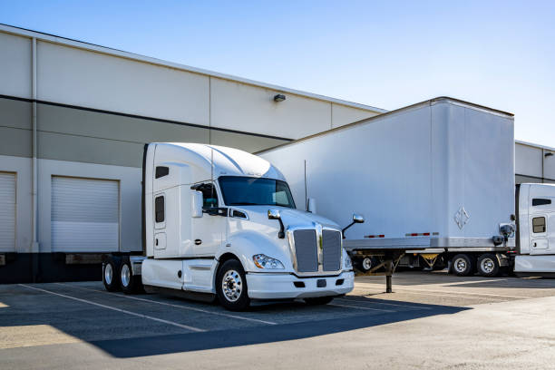 istock Powerful white big rig semi truck tractor standing side by side with loading cargo dry van semi trailer in warehouse dock gates at sunny day 2184489263