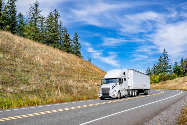 istock Popular comfortable white big rig semi truck transporting cargo in dry van semi trailer running on the winding mountain road between two hills 2184489346