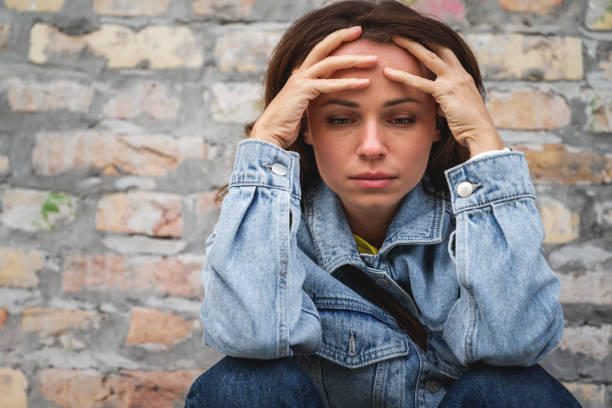 istock Distraught woman with head in hands sitting next to a brick wall outdoors, looking down. 2184495397