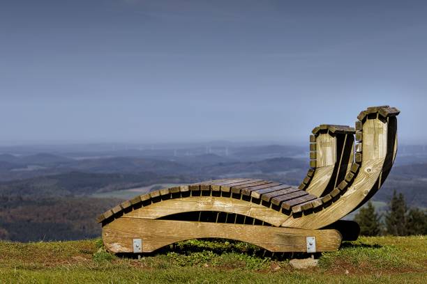 Wooden lounge chair on a hilltop overlooking a vast landscape with clear blue skies, capturing serenity and nature's beauty.