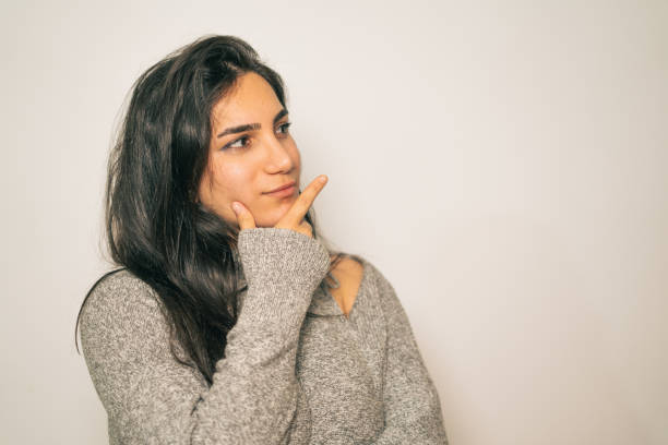 A young woman in a gray sweater strikes a thoughtful pose with a serene expression, set against a minimal indoor background.