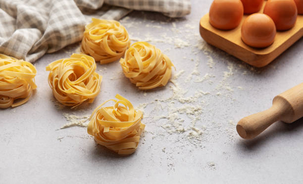 istock Tagliatelle nests with flour, eggs and rolling pin composing a beautiful food background 2184514232