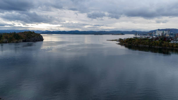 A serene coastal landscape with calm waters, distant hills, and a cloudy sky. The scene includes a small town on the right and lush greenery on the left, creating a peaceful atmosphere.