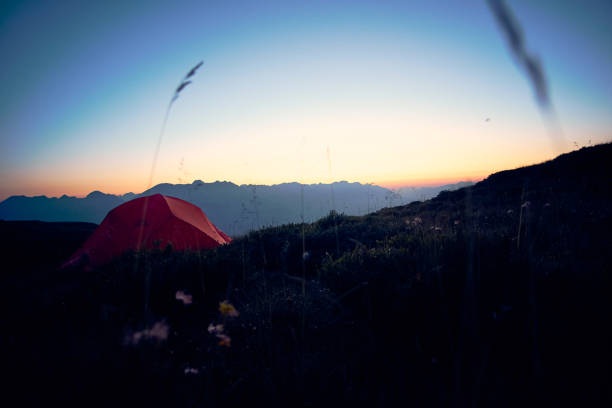 A vibrant red tent sits on a grassy hill as the sun sets behind majestic mountains. The sky is painted with shades of orange and purple, creating a peaceful atmosphere.