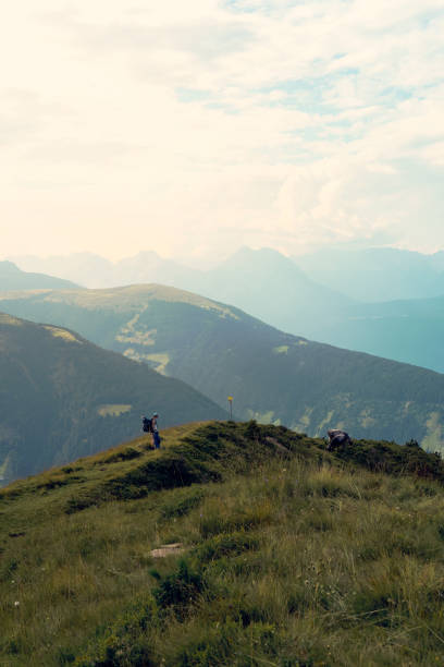 Two hikers traverse a grassy ridge, enjoying breathtaking mountain views under a partly cloudy sky. The sun casts a warm glow on the hills in the distance.