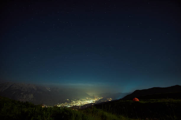 A bright valley glimmers under a starry night sky, with a single tent pitched on the hillside. The peaceful ambiance showcases nature's beauty and tranquility.