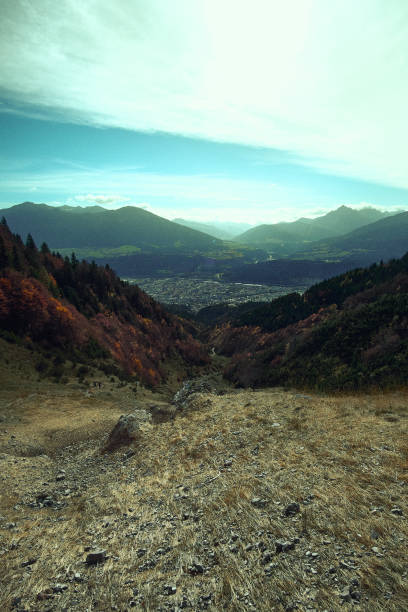 A stunning perspective from a rocky mountain valley reveals a quaint town nestled below. Rolling hills and majestic distant peaks stretch into the horizon under a clear sky.