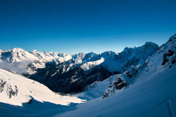 A breathtaking view of snow-covered mountains stretches across the horizon under a clear blue sky. The bright sunlight illuminates the winter landscape, showcasing the beauty of nature.